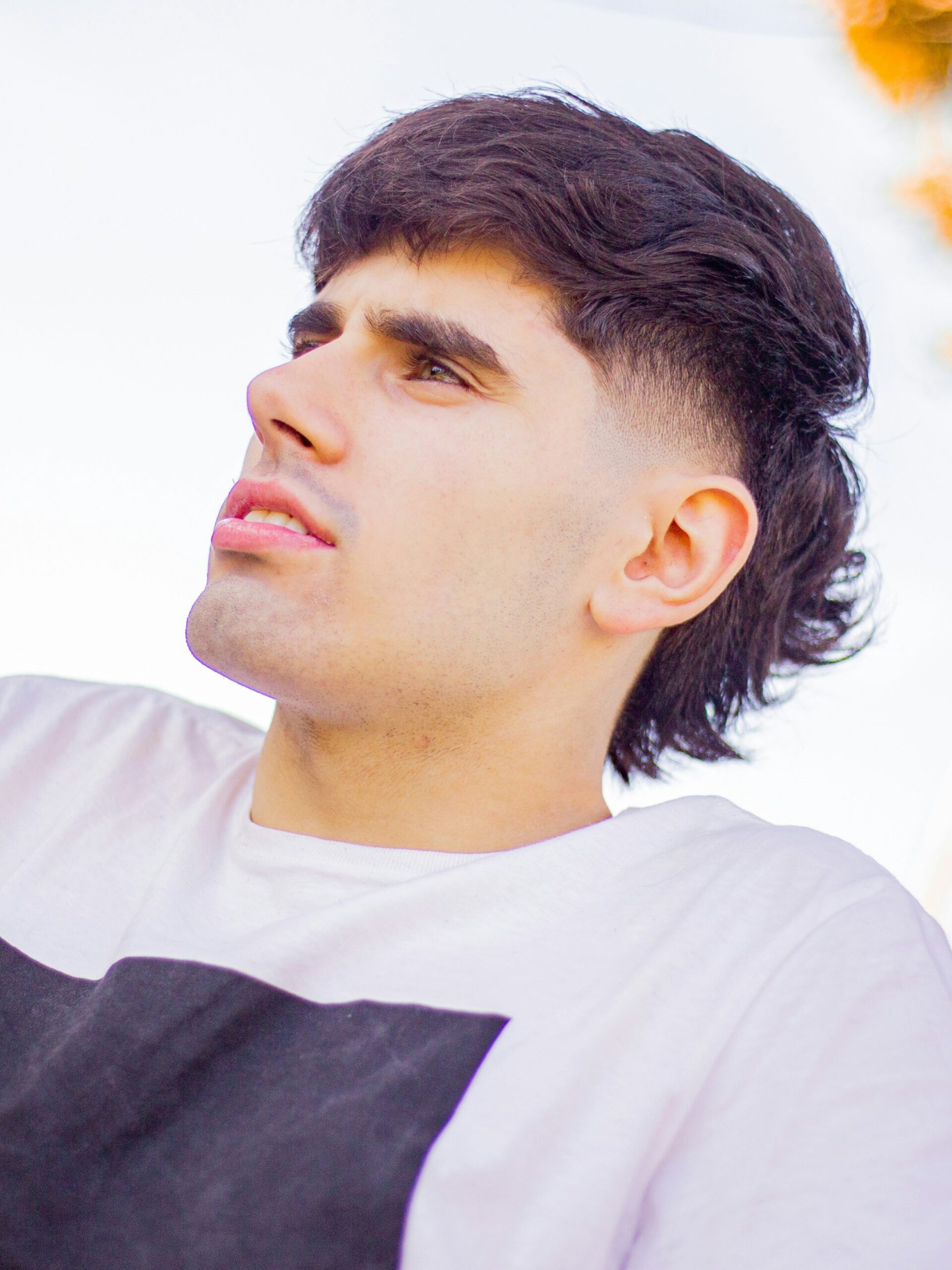 Een jonge man met een strak fade-mullet-kapsel in een wit T-shirt, zittend langs de weg op een zonnige dag.