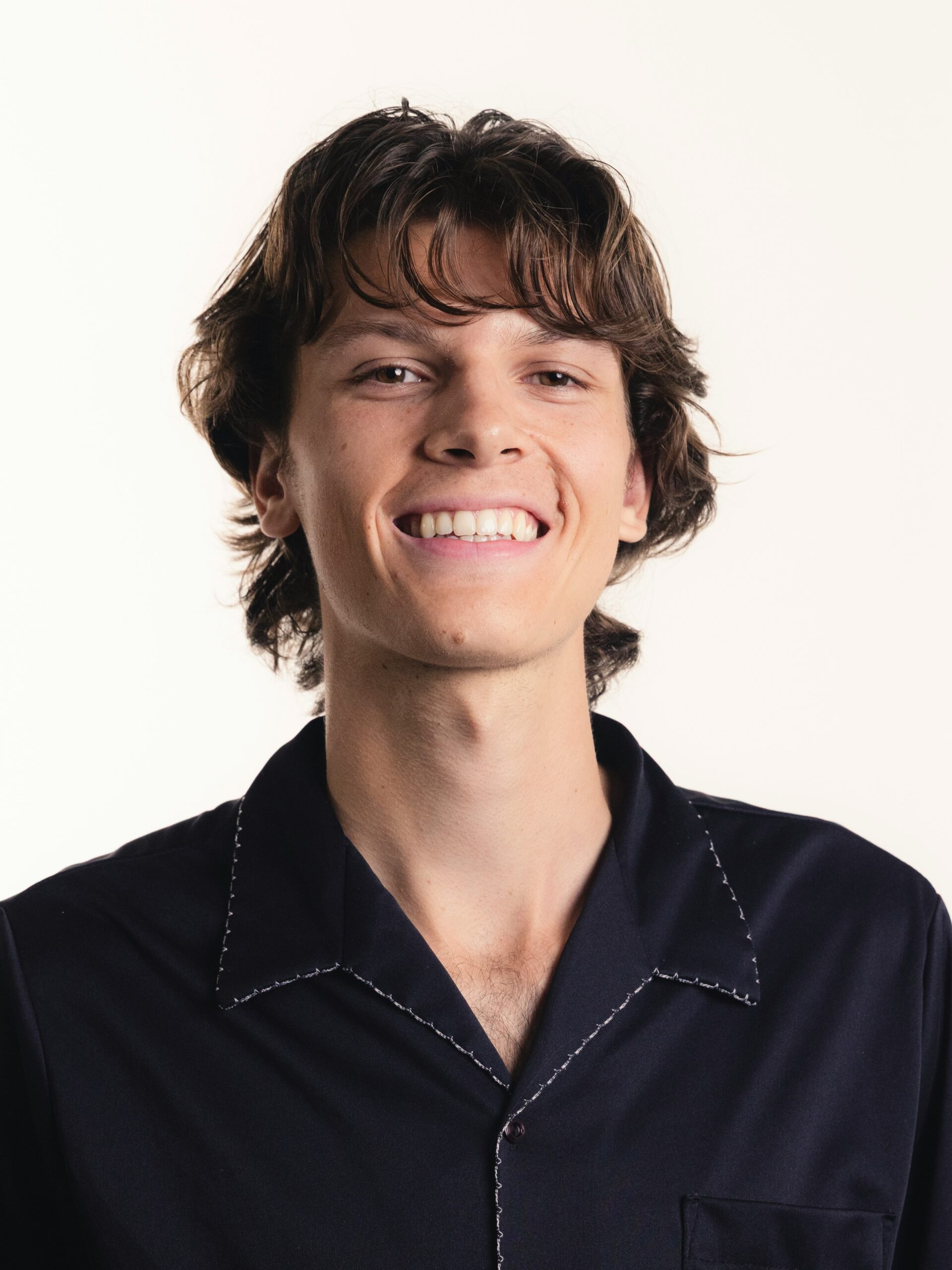 Een jonge man met een gelaagd, gestructureerd mullet-kapsel in een donkerblauw overhemd, lachend.