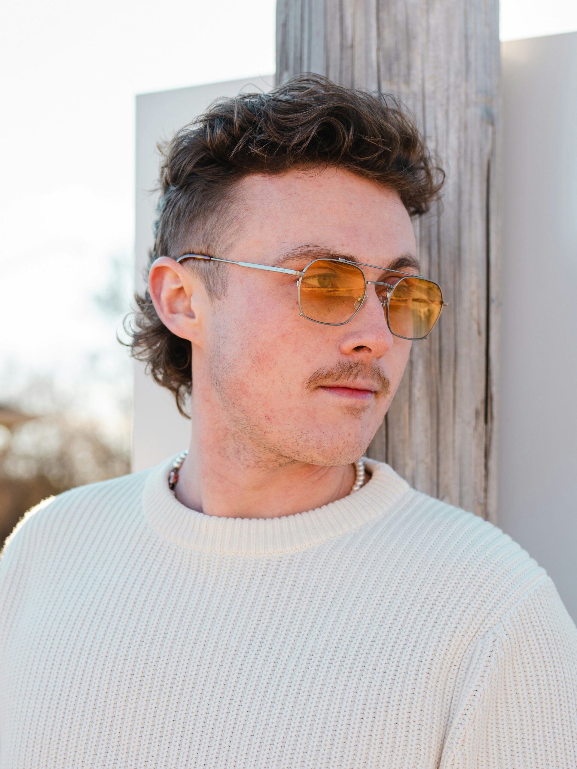 Een jonge man met een krullend mullet-kapsel leunend tegen een muur buiten op een zonnige dag.