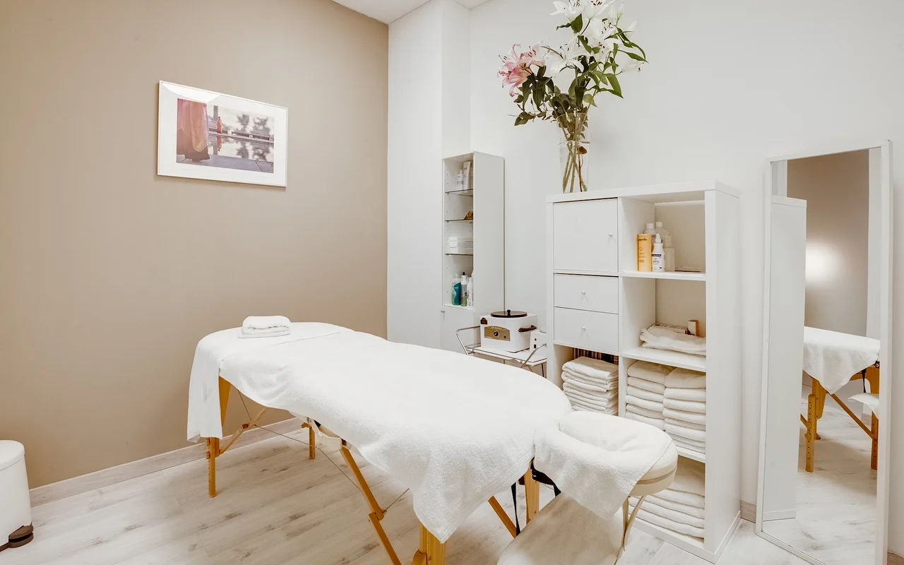 Een serene behandelkamer met een witte massagetafel gedrapeerd met schone handdoeken. De ruimte heeft neutrale beige en witte wanden, een witte opbergkast met gevouwen handdoeken, en een hoge glazen vaas met witte en roze lelies.