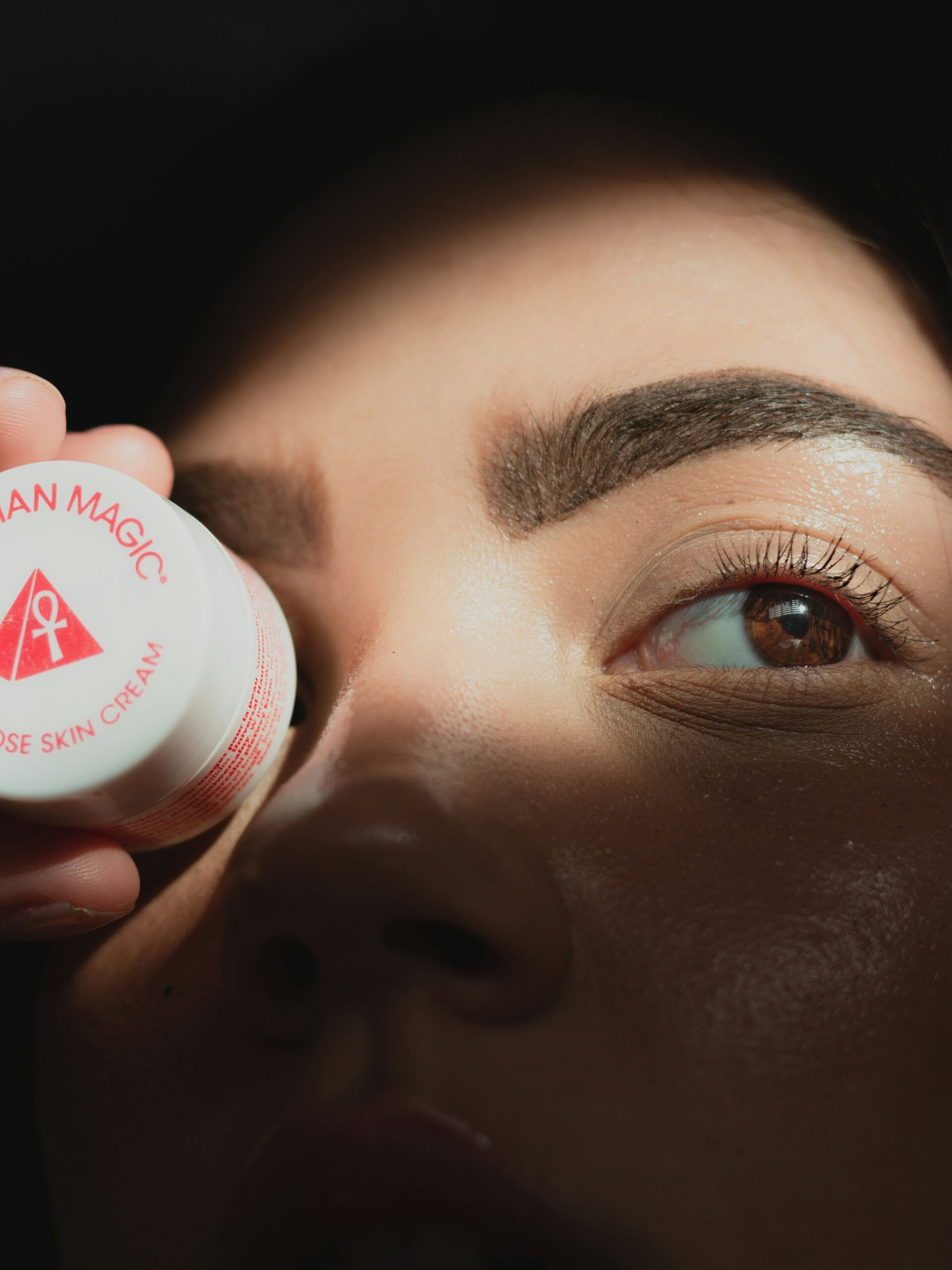 Close-up van het gezicht van een vrouw met microblading op haar wenkbrauwen, die een klein rond potje crème voor één oog houdt