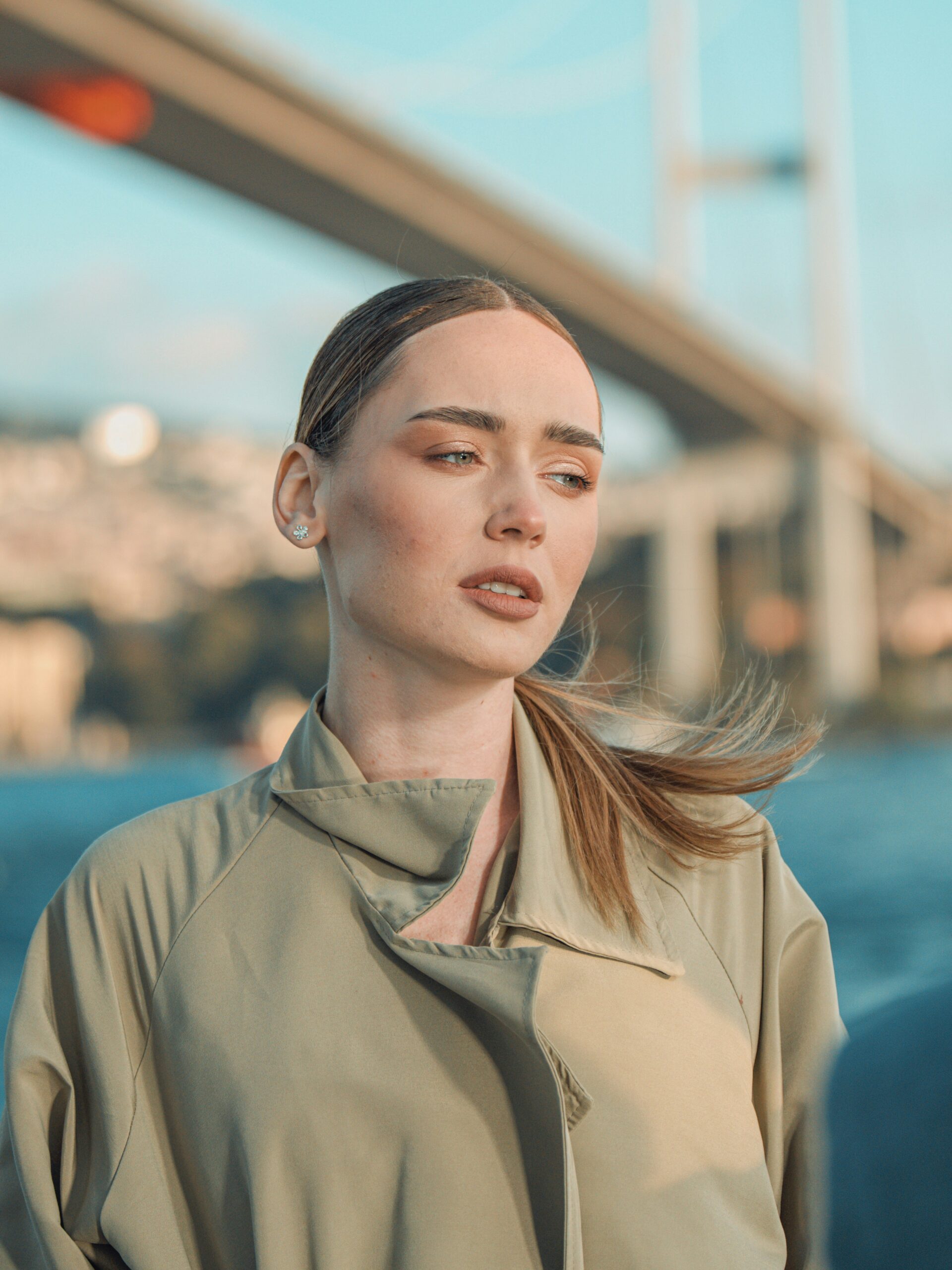 Een vrouw met microblading op haar wenkbrauwen en blond haar dat naar achteren is gebonden, in een trenchcoat, staand voor een brug in Istanbul