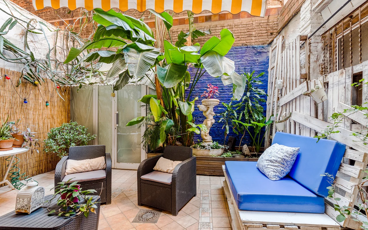 Een eclectisch buitenterras of wachtruimte met een levendig blauwe accentmuur en weelderige tropische planten. Er staan donkere rieten fauteuils, een felblauwe bank met kussens, en een geel-wit gestreepte luifel
