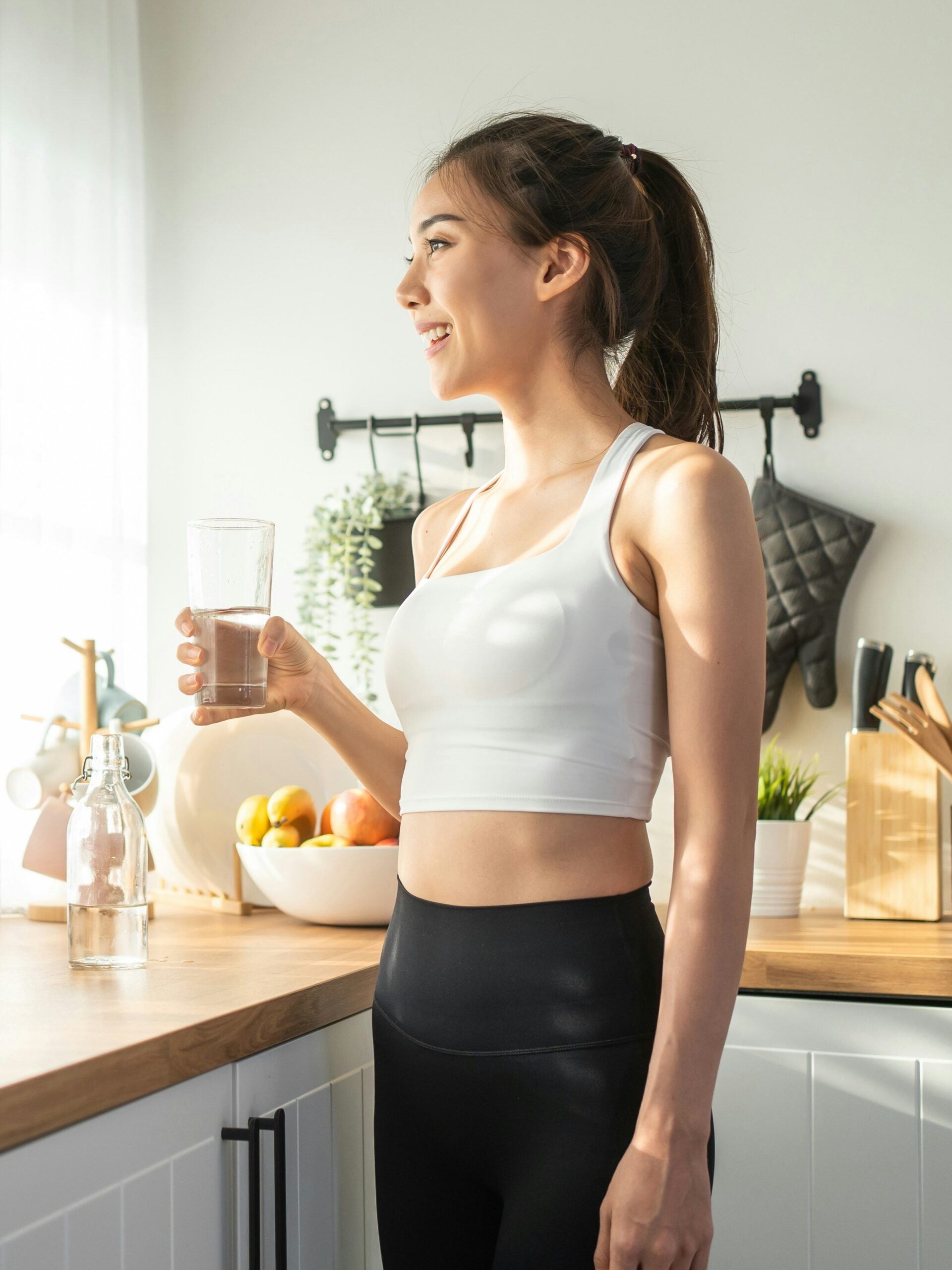 Zijaanzicht van een vrouw die water drinkt in haar keuken voor een zonnig raam