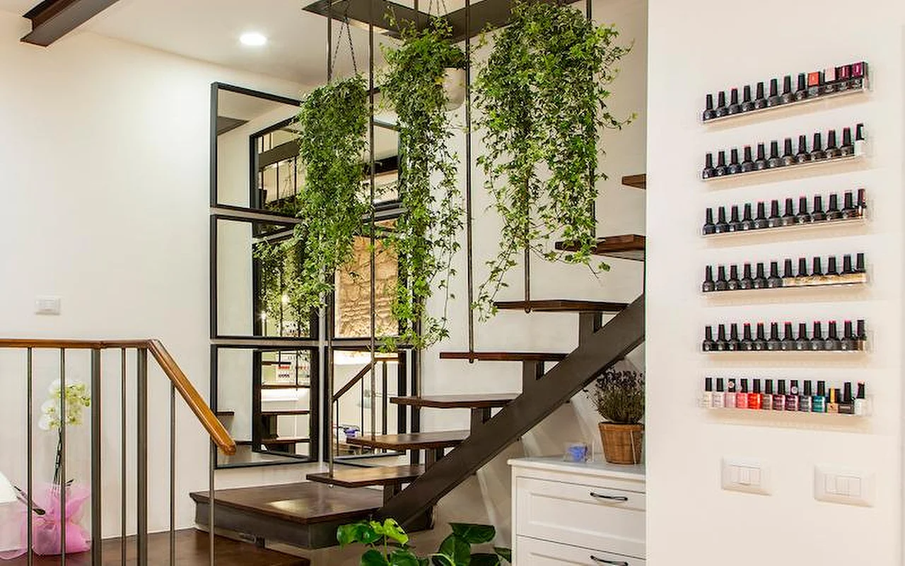 Een stijlvol modern interieur met een vrijdragende trap van hout en metaal. Weelderige hangplanten hangen tussen de treden aan het plafond, en rijen kleurrijke nagellakflesjes staan uitgestald op slanke transparante schappen aan de witte muur.