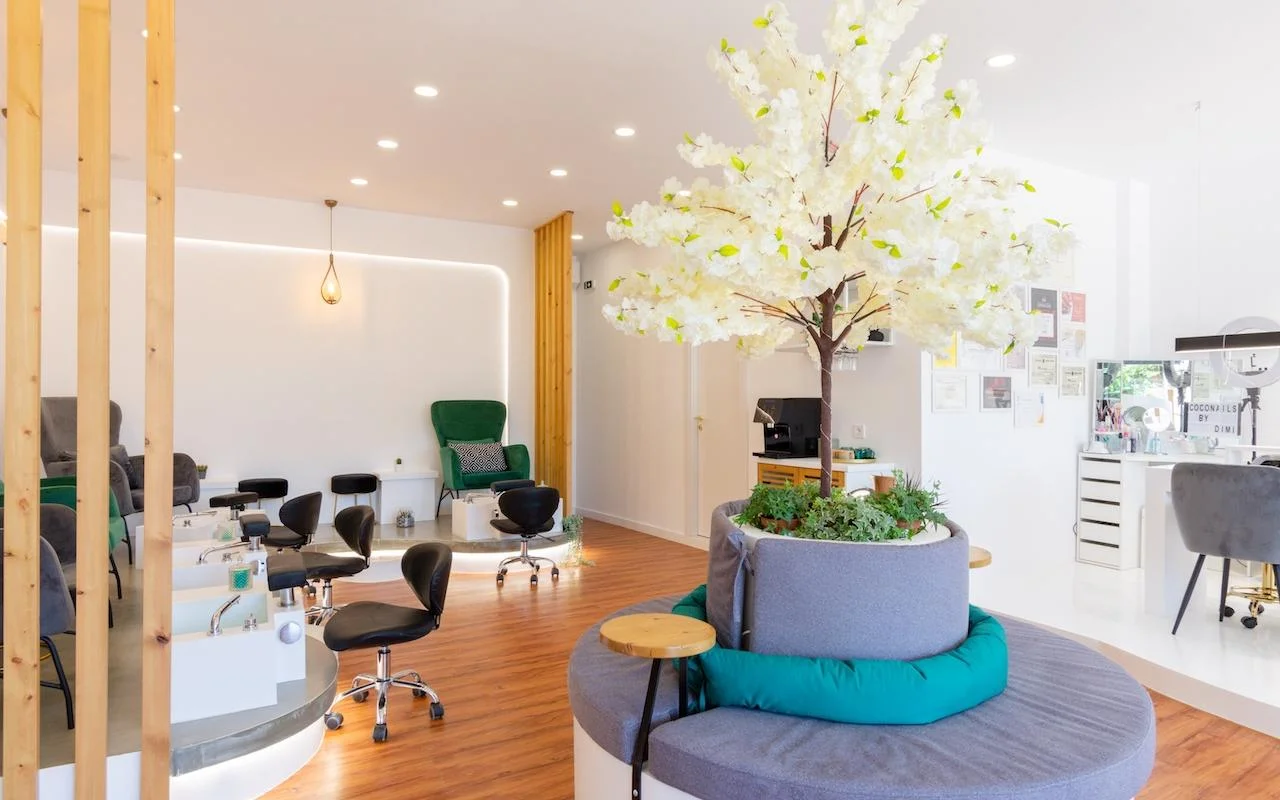 Een open, door de natuur geïnspireerde nagelsalon met een grote decoratieve witte bloesemboom als middelpunt, smaragdgroene fluwelen fauteuils bij pedicurestations, houten latscheidingswanden en een warme houten vloer.