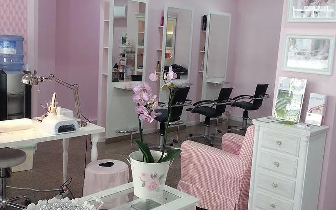 Het interieur van een lichte, roze-thema haar- en nagelsalon. Links een witte manicurestation met bureaulamp en nageldroogapparaat. In het midden een orchidee in een witte pot op een salontafel naast een roze-wit geruite fauteuil. Op de achtergrond meerdere kappersstations met hoge spiegels en zwarte kappersstoelen tegen lichtroze muren.