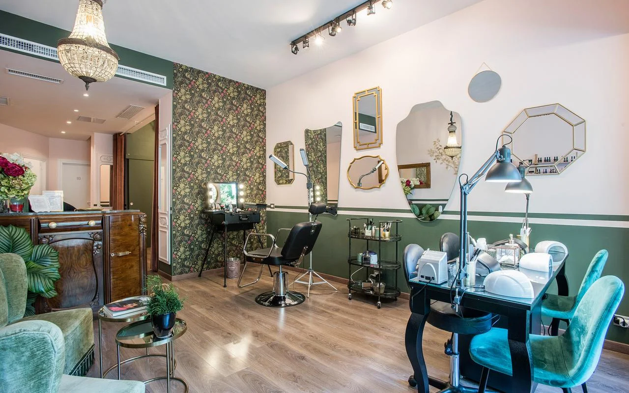 Een salon met vintage-chic uitstraling: witte en bosgroene muren, bloemenbehang, een verzameling onregelmatig gevormde goudkleurige spiegels, een professioneel make-up station en weelderige groene fluwelen zitjes.