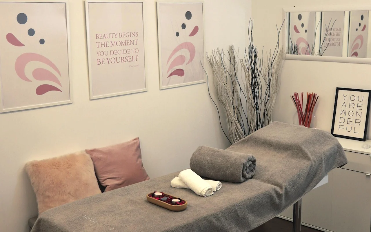 Een lichte behandelkamer met een massagetafel bedekt met een grijze handdoek. De muur is versierd met abstracte kunst in pastelkleuren en motiverende quotes, terwijl decoratieve witte takken en kaarsen een kalme sfeer creëren.