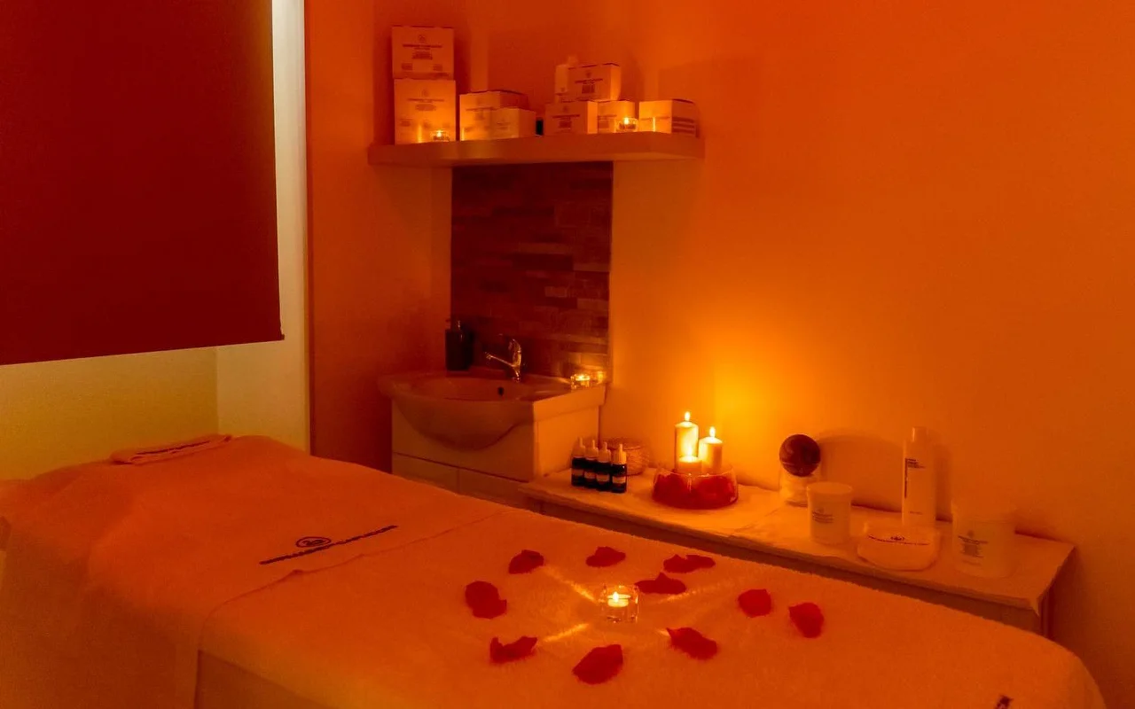 Een massageruimte met warme, sfeervolle oranje verlichting en flakkerende kaarsen. Rode rozenblaadjes liggen in een cirkelvormig patroon rond een centrale kaars op de behandeltafel, met beautyproducten netjes georganiseerd op een plank erachter.