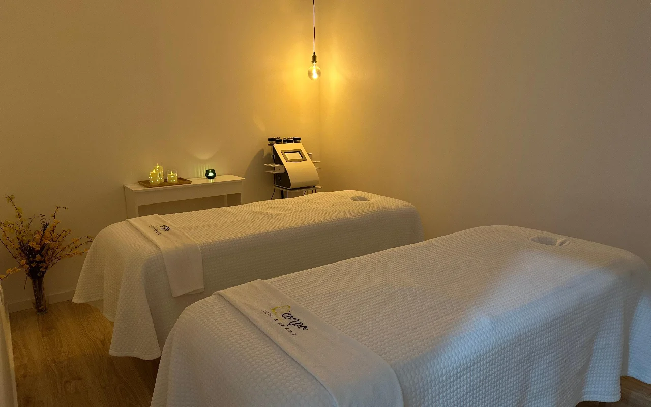 Een rustige massage- of behandelruimte met twee behandeltafels naast elkaar, opgemaakt met witte handdoeken en lakens. De ruimte wordt verlicht door een enkele warme, onbedekte hanglamp en kleine kaarsjes op een bijzettafel, wat een serene sfeer creëert. Een gespecialiseerd cosmetisch apparaat staat op de achtergrond tussen de bedden.