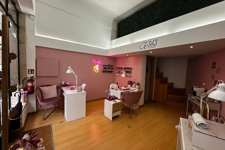 Een stijlvolle nagelstudio met lichtroze muren en warme houten vloeren. De ruimte bevat meerdere witte manicuretafels met oud-roze fluwelen stoelen. Een felle roze neonreclame in de vorm van een hand met nagellakflesje en het woord 'Nails' gloeit aan de muur, en rijen nagellak staan uitgestald op kleine planken. Een mezzanine met een groene kunsthegwand is zichtbaar boven.