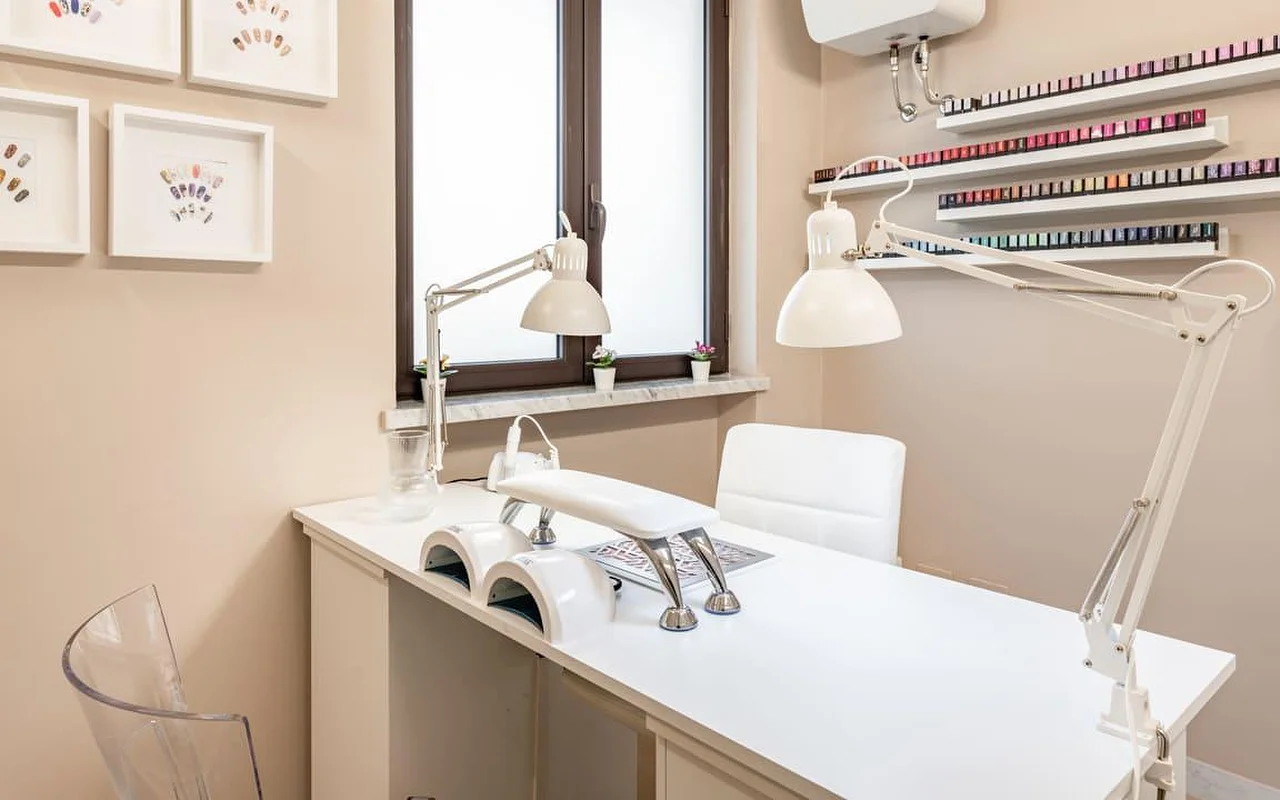 Een nagelstation bij AG The Beauty Room's met een schoon wit bureau, dubbele uv-lampen, verstelbare werklampen en schappen vol rijen gekleurde nagellak. Aan de warmgetinte wanden hangen ingelijste nageldesigns.