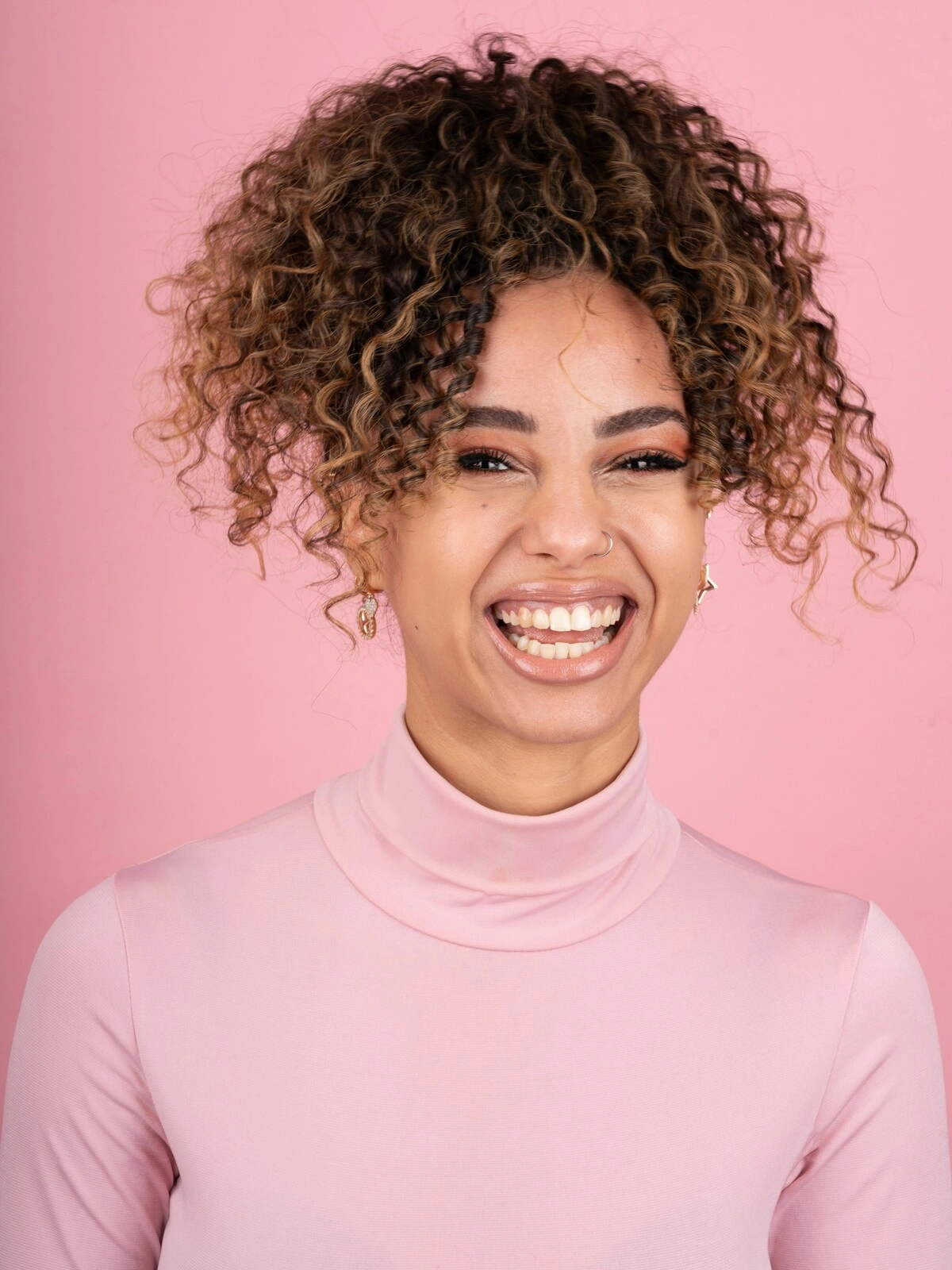 Studiofoto van een lachende vrouw met balayage op afrohaar, gekleed in een zachtroze coltrui