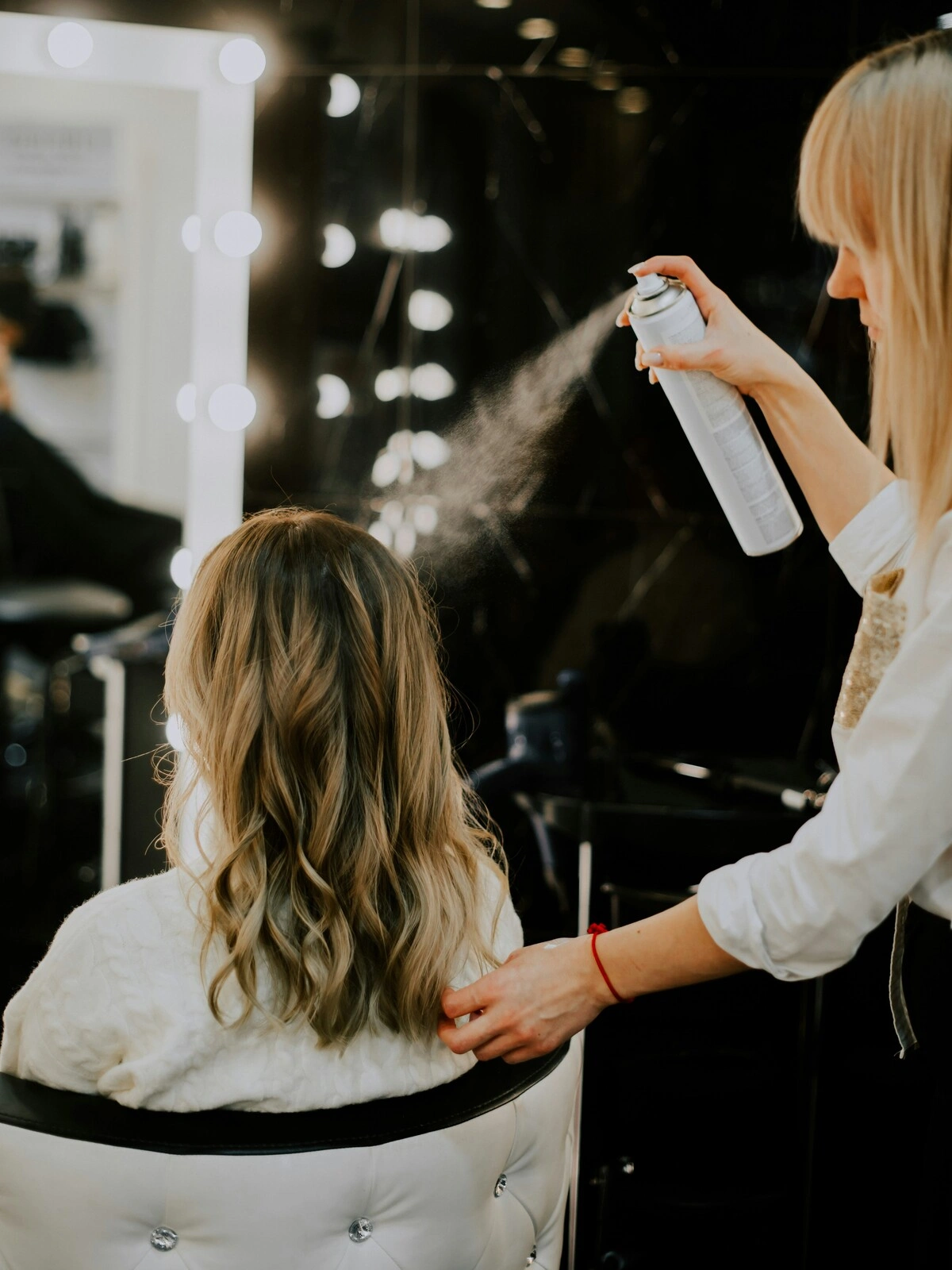 Een kapster spuit een finishingproduct op het haar van een klant na een balayage, föhnbeurt en het in golven stylen