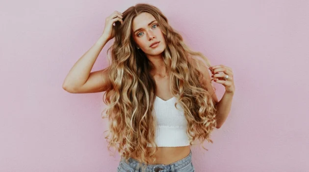 Studiofoto van een vrouw met heel lang, golvend, door de zon gekust balayage-haar, gekleed in een lichtblauwe jeans en een wit kort gebreid topje