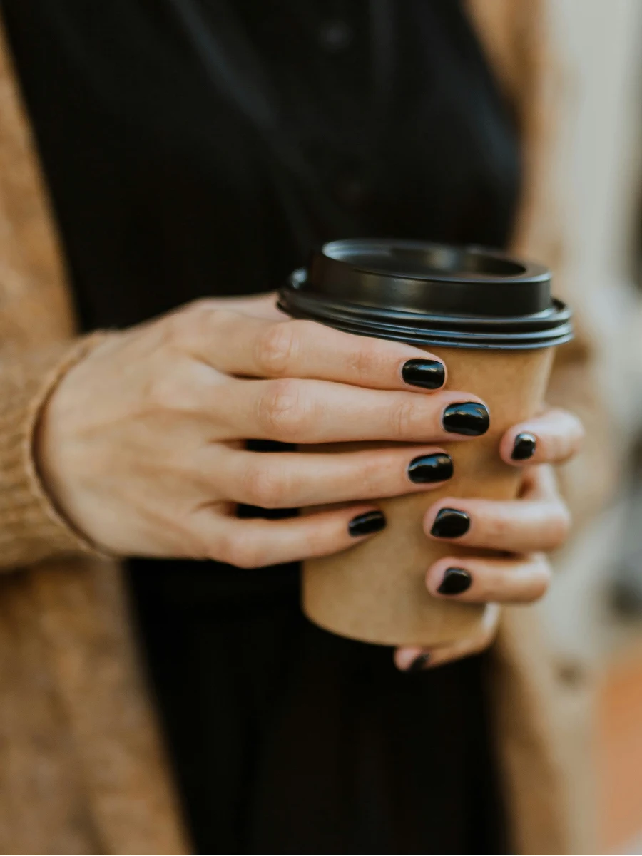 Close-up van een vrouw met een glanzende zwarte gelmanicure die een koffiebeker om mee te nemen vasthoudt