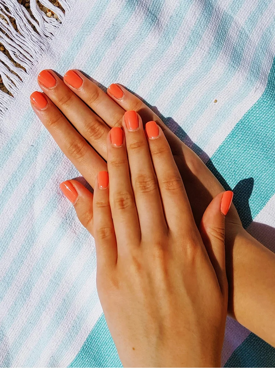 Bovenaanzicht van twee handen met een glanzende oranje gelmanicure, rustend op een blauw-wit gestreepte strandhanddoek