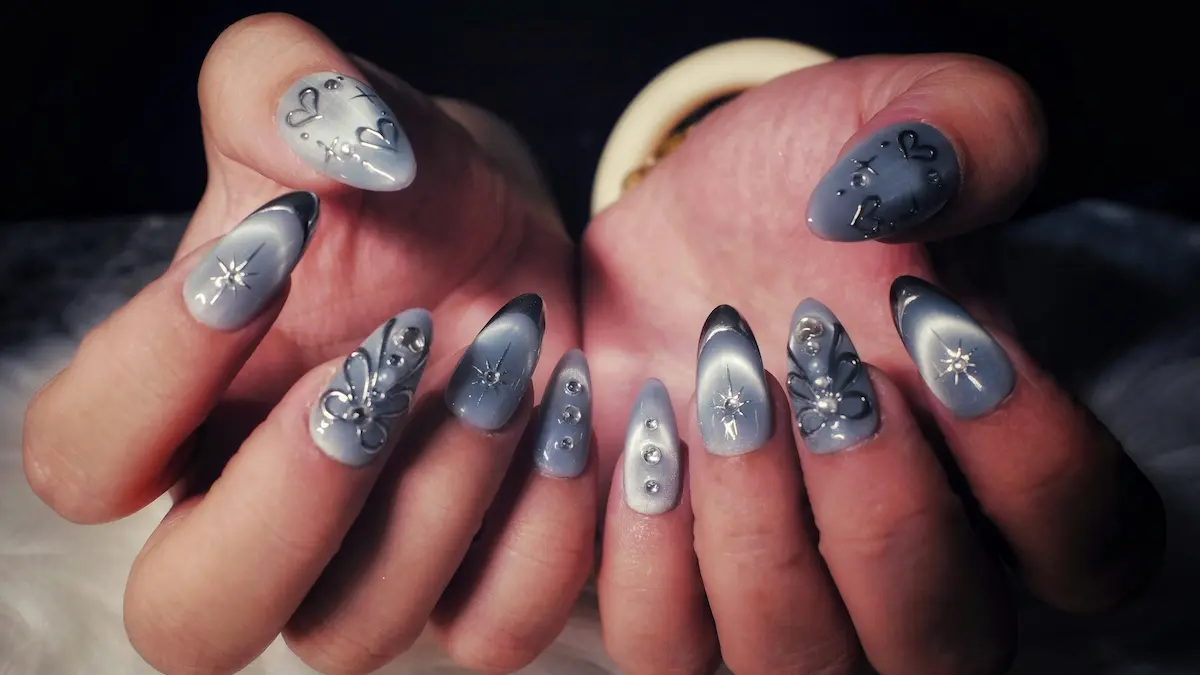 Close-up van de handen van een vrouw met gedetailleerde acrylnagels met een grijs cat-eye-design, zilveren hartjes en sterren en sprankelende steentjes