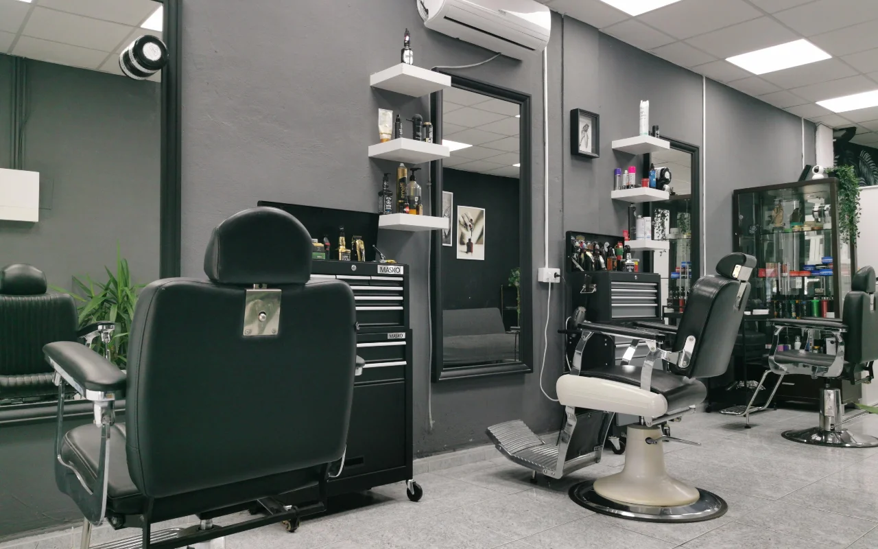 Een traditionele barbershop met houtskoolgrijze muren, klassieke zwartleren barbierstoelen met chromen voetstuk en voetsteun, witte zwevende planken vol verzorgingsproducten en grote spiegels op een betegelde vloer.