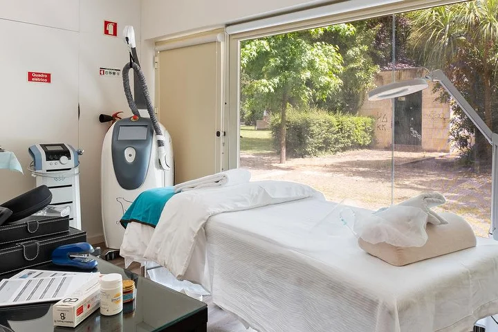 Een behandelkamer met een wit gedrapeerde behandeltafel naast een raam van vloer tot plafond op een groene tuin, een apparaat voor lichaamsbehandelingen op een statief, een vergrootlamp en een glazen bijzettafel met producten.