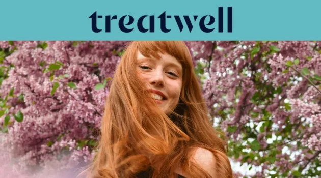 Close-up portret van een glimlachende jonge roodharige vrouw met een pony, wiens haar in de wind waait voor een achtergrond van roze seringentakken. Een lichtblauwe banner bovenaan toont het "Treatwell"-logo in het donkerblauw.