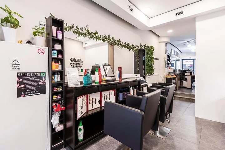 Een smalle kapsalon met zwarte stylingstoelen langs een spiegelwand, een donkere plank vol haarverzorgingsproducten, een groene slinger die over de spiegel valt en een ingelijst Made In Brazil-bord op een witte zuil.