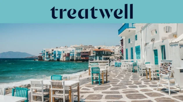 De boulevard van Klein Venetië in Mykonos-stad: witgekalkte huizen met kleurige houten balkons aan het water, daarachter de turkooizen Egeïsche Zee en op de voorgrond witte en petrolblauwe cafétafels op een geplaveid terras.