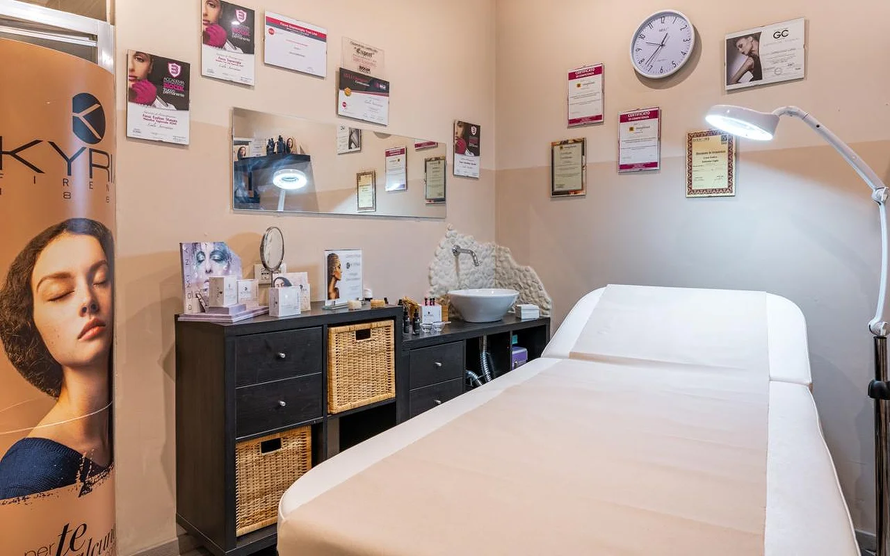 Een behandelkamer bij Oltre la bellezza met een witte behandeltafel, een verstelbare vergrootlamp en wanden vol professionele certificaten en diploma's. Onder een spiegel staan donkere rieten opbergkasten, met verzorgingsproducten op het werkblad.