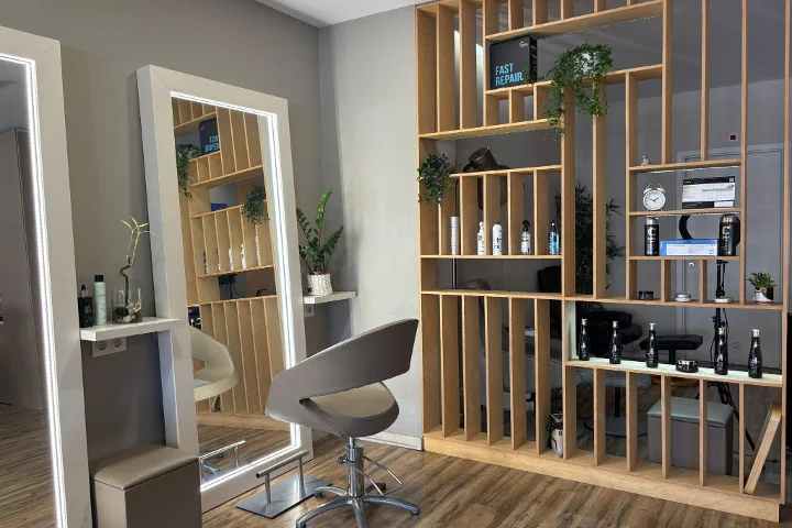 Een kappersplek met een grijze stylingstoel voor een ingelijste, met LED verlichte spiegel, een schap-scheidingswand van houten latten met stylingproducten, een klok en planten, op een vloer met houtlook.