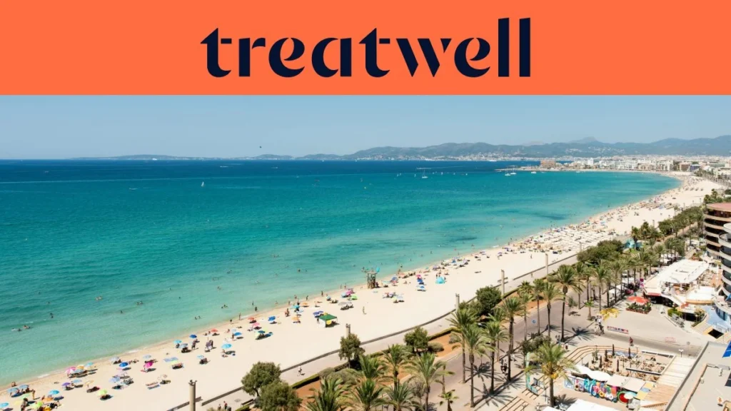 Een luchtfoto van Playa de Palma op Mallorca, met een lange boog van goudkleurig zand, rijen parasols, kalm turquoise water en een met palmen omzoomde boulevard langs de stad, met heuvels in de verte onder een strakblauwe lucht.