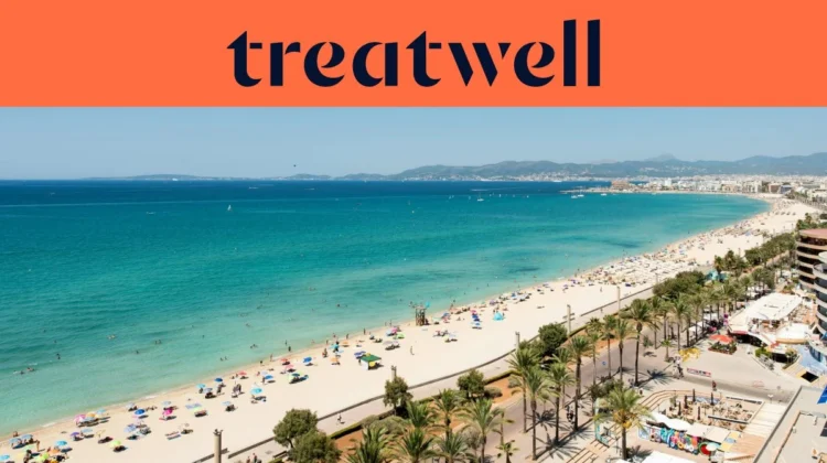 Een luchtfoto van Playa de Palma op Mallorca, met een lange boog van goudkleurig zand, rijen parasols, kalm turquoise water en een met palmen omzoomde boulevard langs de stad, met heuvels in de verte onder een strakblauwe lucht.