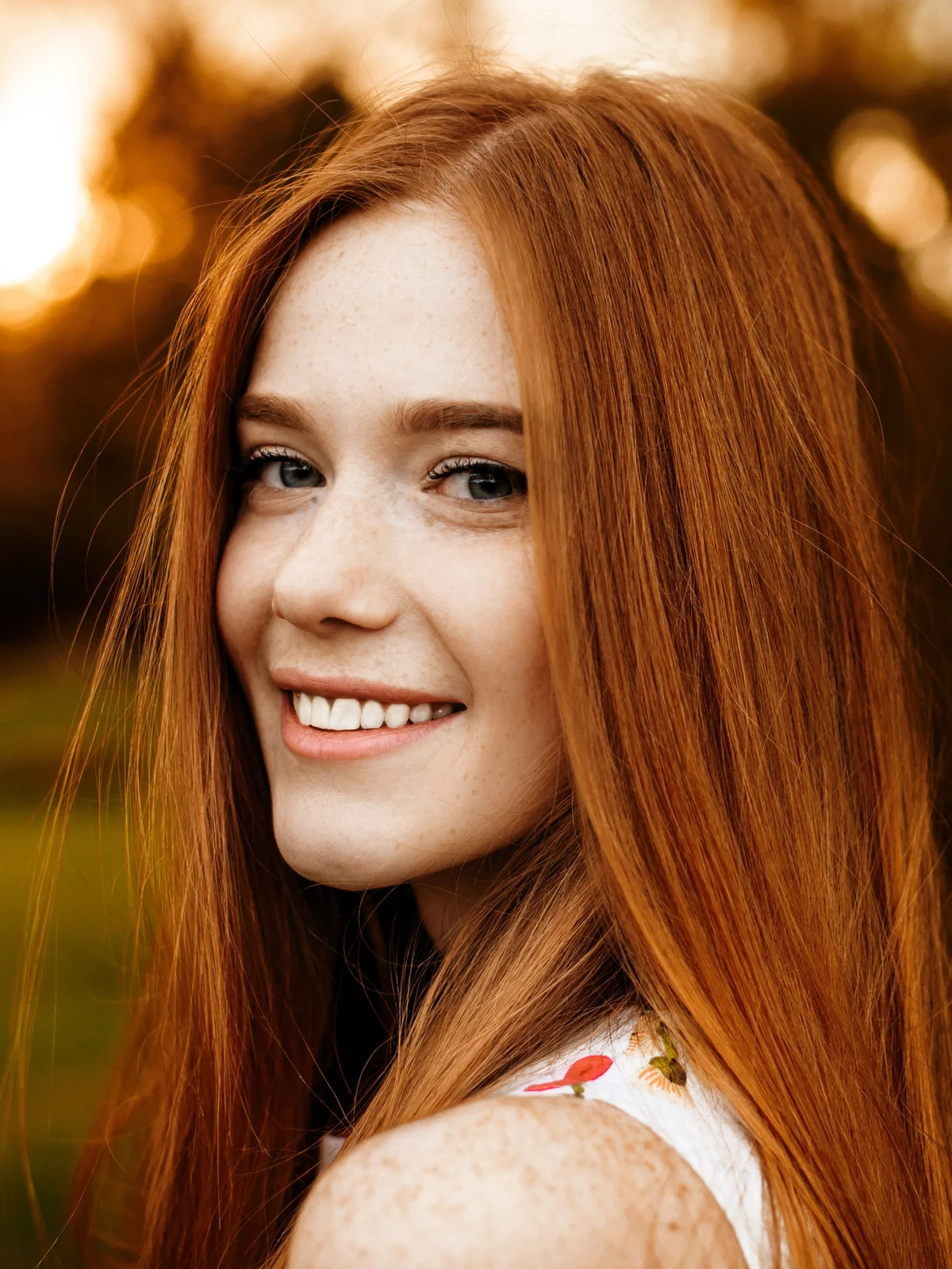Een vrouw met lang rood haar en nette, gedefinieerde wenkbrauwen glimlacht terwijl ze buiten in de natuur wandelt in het afnemende daglicht