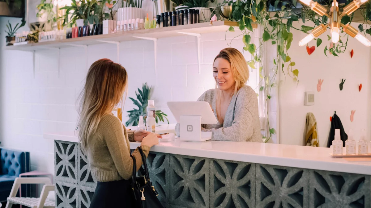 Een vrouw staat aan een salonbalie terwijl een receptioniste haar volgende behandeling boekt; de salon is netjes, met een witte balie, opgeruimde schappen met producten en planten en bloemen als decoratie