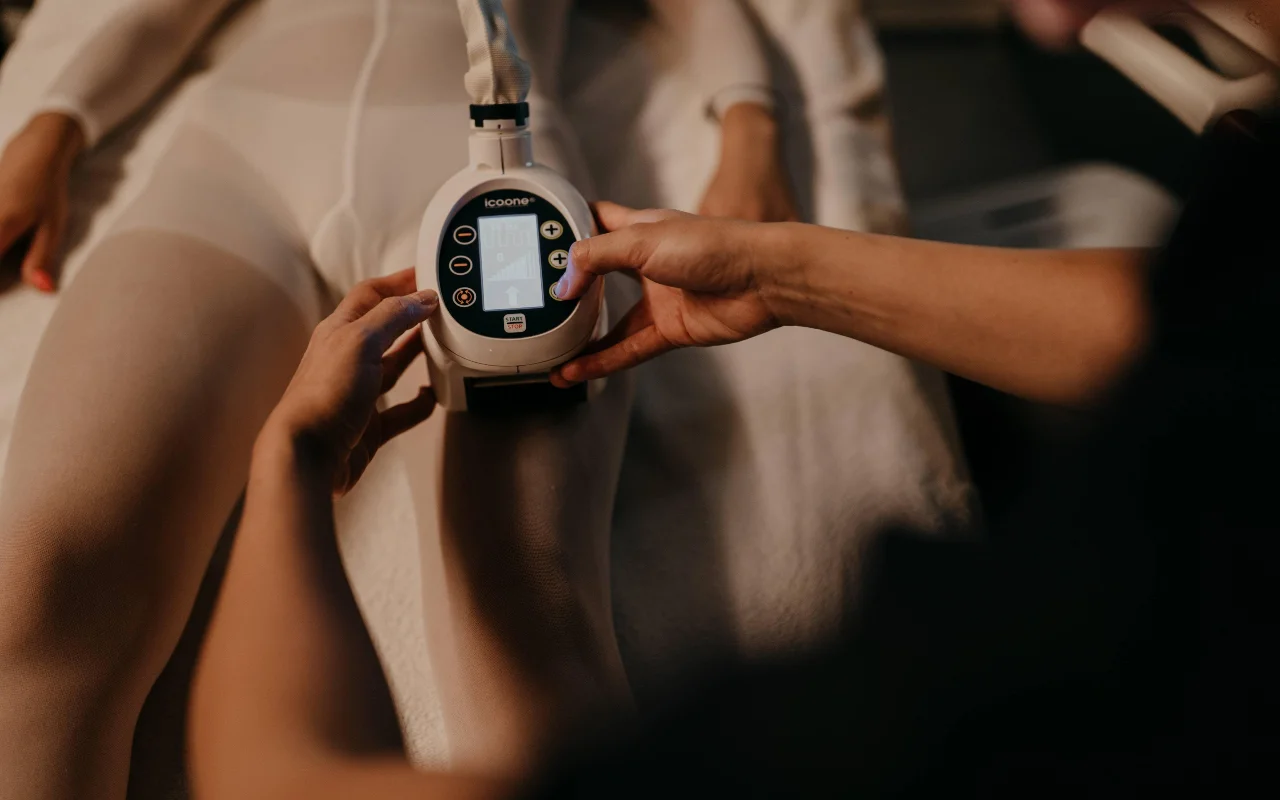 Een body-contouringbehandeling bij Beauty Room Romina Bongiovanni, met een schoonheidsspecialiste die een icoone-apparaat tegen het been van een klant in een wit behandelpak houdt, in warm, gedempt licht.