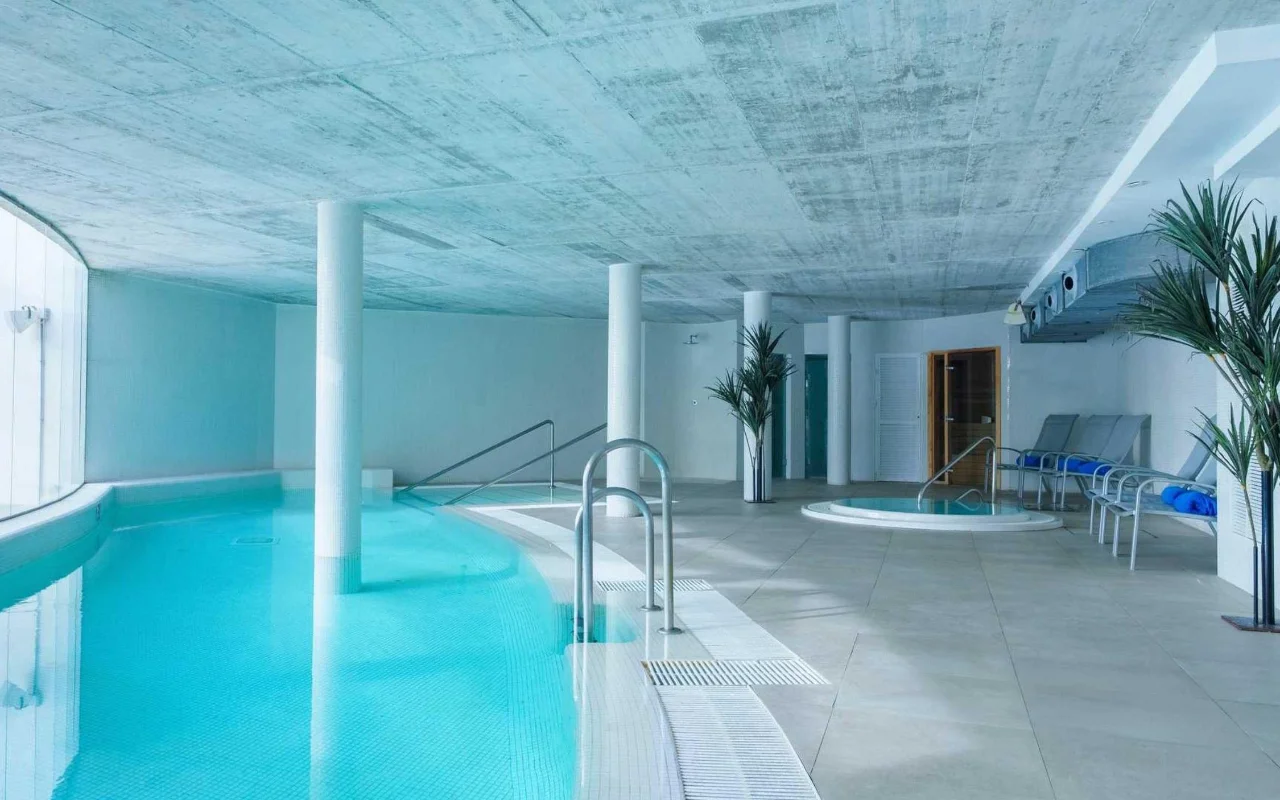 Het overdekte spazwembad van Benalmádena Beach, met een gebogen turquoise bad, een betonnen plafond, een kleine jacuzzi en ligbedden tussen hoge palmen in potten.