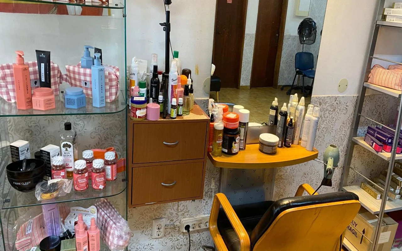 De werkplek van een kapper met een wandspiegel en een houten werkblad vol stylingproducten, een glazen vitrine met haarverzorgingsproducten en supplementen ernaast, en een klassieke kappersstoel in hout en zwart.