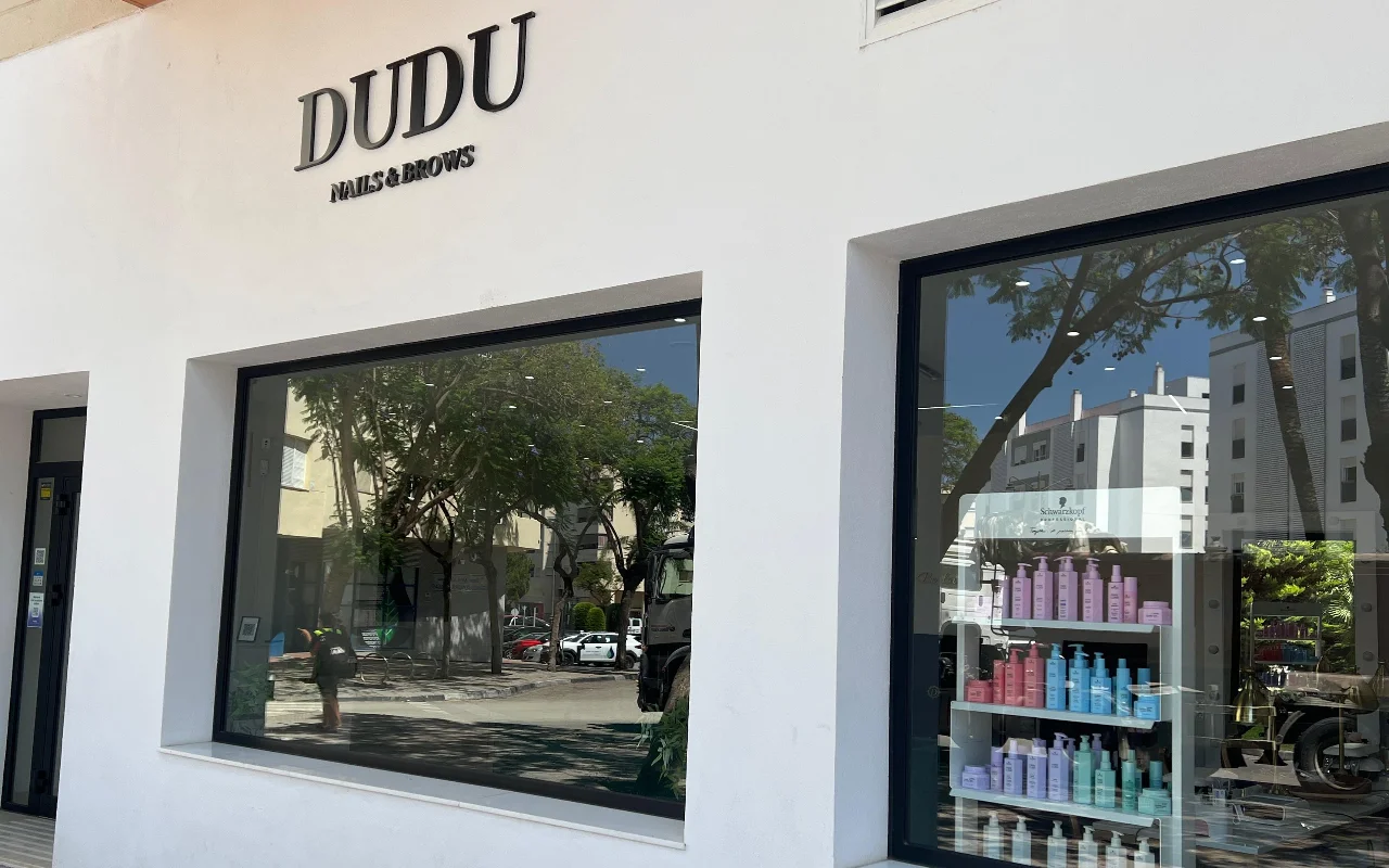 De witte gevel van Dudu Nails and Brows in Estepona, met de naam in donkere letters boven brede etalages vol rijen haarproducten.