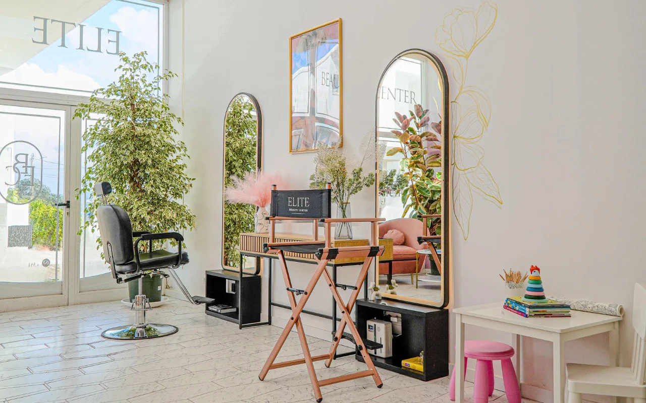 Een lichte, luchtige, helemaal witte beautysalon met gewelfde goudomlijste spiegels, een zwarte kappersstoel en een opklapbare regisseursstoel, hoge groene planten, roze accenten en een klein kinderhoekje bij het raam.