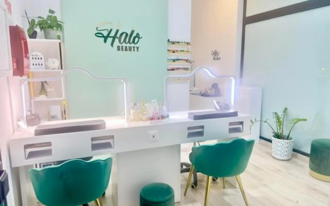 Een nagelstudio met een muntgroene accentmuur met het Halo Beauty-logo, een witte dubbele manicurebalie met verlichte plexiglas schotjes, twee smaragdgroene fluwelen kuipstoeltjes, een wandrek vol nagellak en potplanten.