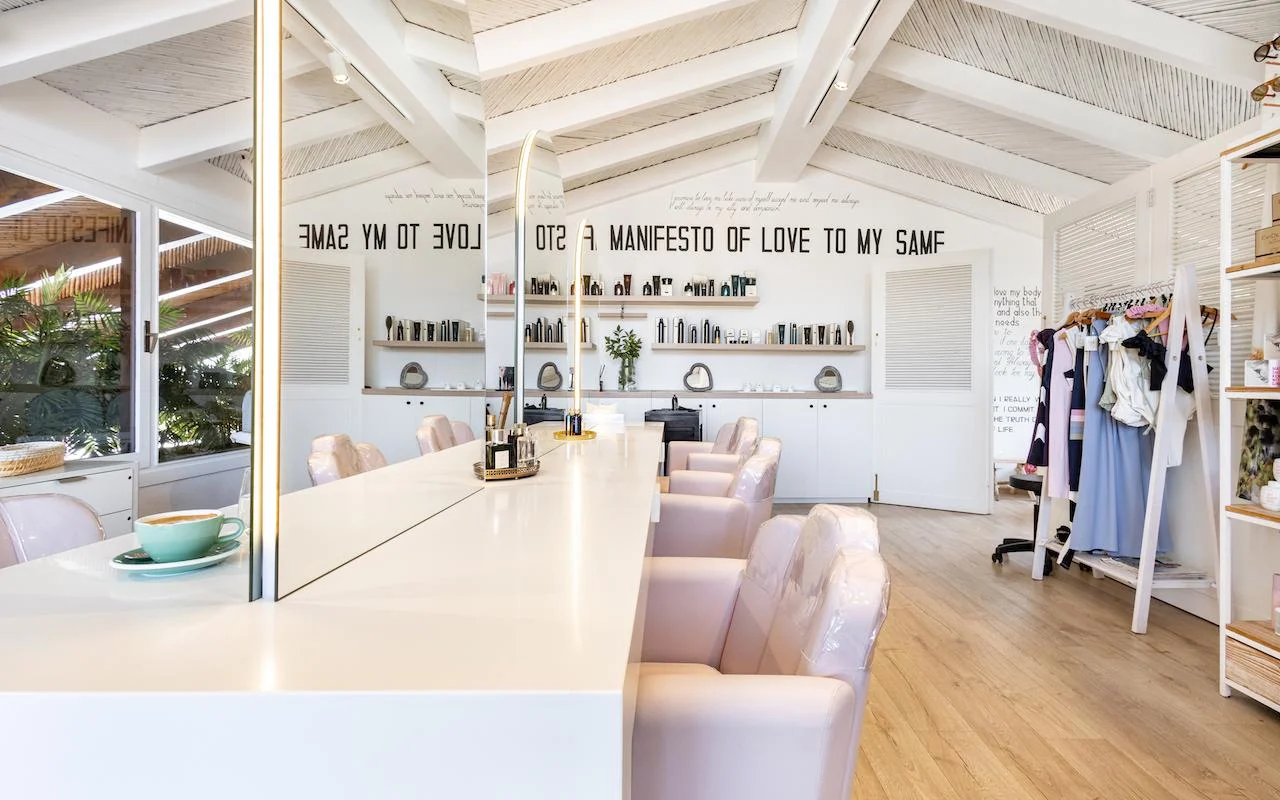 Het lichte interieur van Milk & Roses in Puente Romano, Marbella, met een witte stylingbalie, lichtroze stoelen, een balkenplafond en productplanken langs de muur.