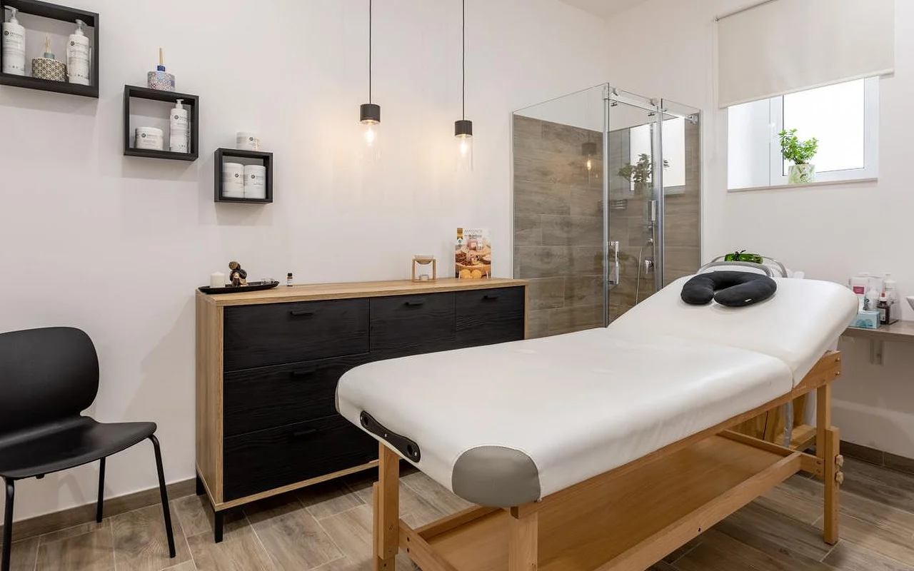 Een massageruimte bij Thea Estetica, met een houten massagetafel met zwarte gezichtssteun, een ladekast in zwart en hout, twee hanglampen, kubusvormige wandrekken met producten en een glazen douchecabine met tegels in houtlook.