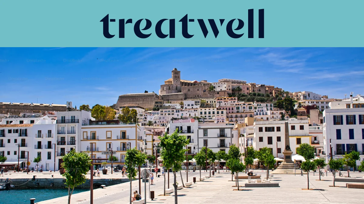 Zonnig uitzicht over Ibiza-Stad, met witgekalkte gebouwen die oplopen naar de kathedraal en de oude vestingmuren van Dalt Vila op de heuvel. Een door bomen omzoomde havenpromenade loopt door de voorgrond onder een strakblauwe hemel.