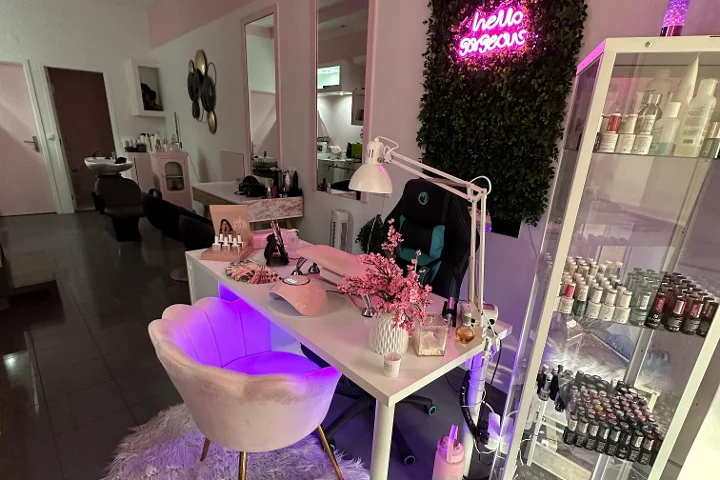 Roze verlicht beautyplekje met een 'hello gorgeous'-neonbord op een groene wand, een crèmekleurige schelpvormige stoel, een witte tafel met kwasten en kersenbloesems, en een vitrine vol kleurige nagellak.