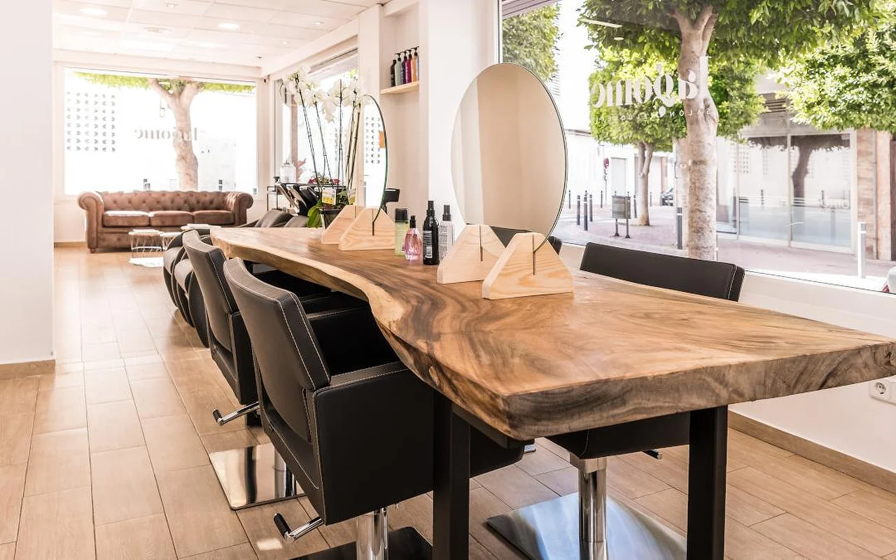 Een lichte kapsalon gebouwd rond een lange houten toonbank met natuurlijke randen, bekleed met zwarte kappersstoelen. Een ronde spiegel en houten productstandaards staan op de toonbank, een bruine leren Chesterfield-bank wacht achterin, en hoge ramen kijken uit op een groene straat.
