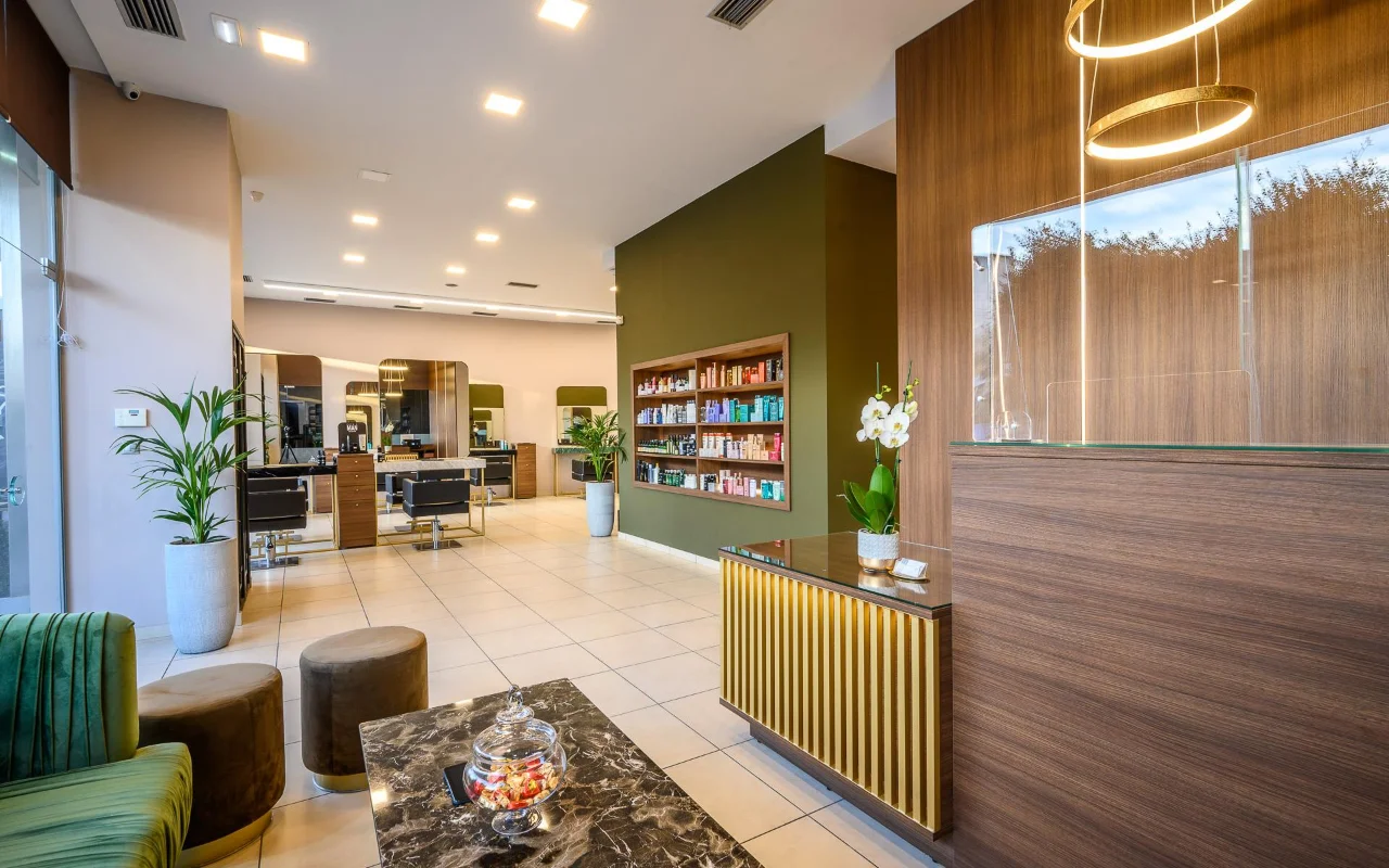 Een chique kapsalon met warme houten lambrisering, een olijfgroene accentwand met productplanken, styling-plekken met grote spiegels, een receptiebalie met gouden lamellen en groene fluwelen zitplaatsen onder ronde gouden hanglampen.