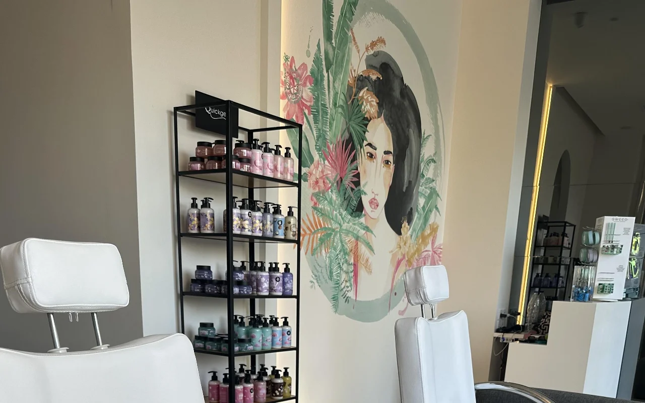 Een studiohoek met twee witte verstelbare behandelstoelen, een zwarte productplank, een van achter verlichte spiegel, en een florale muurschildering van een vrouwengezicht omlijst door tropisch loof.
