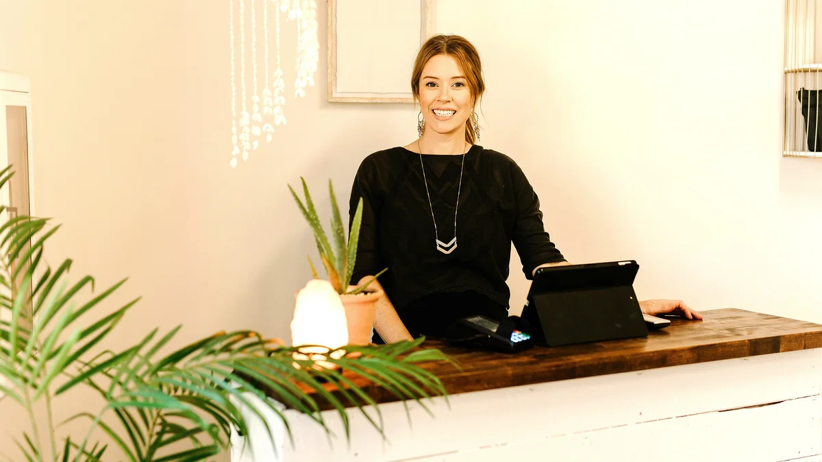 Een lachende vrouw staat achter de receptiebalie van een schoonheidssalon