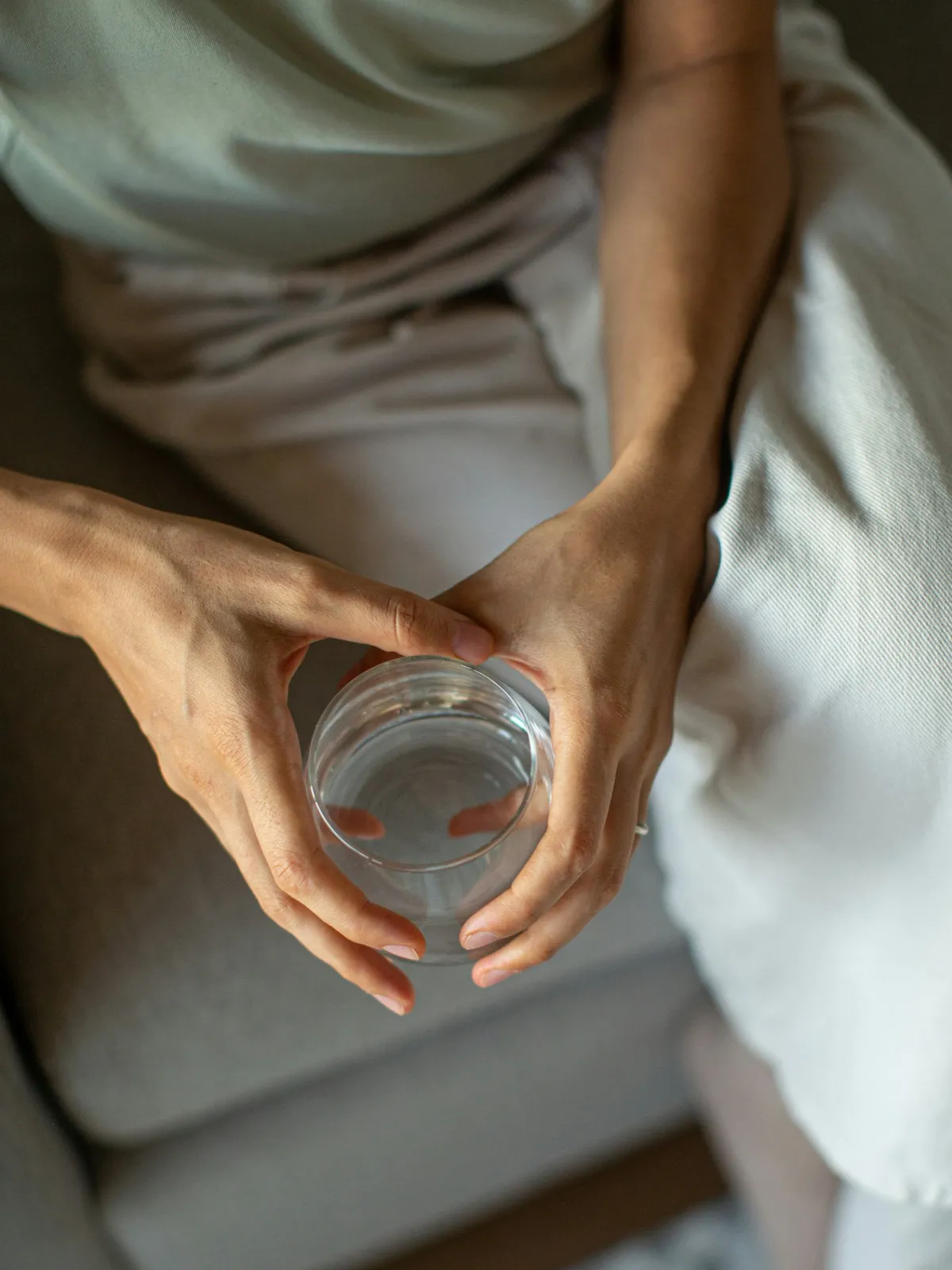 Bovenaanzicht van iemand in losse kleding op een grijze bank, met een glas water in beide handen
